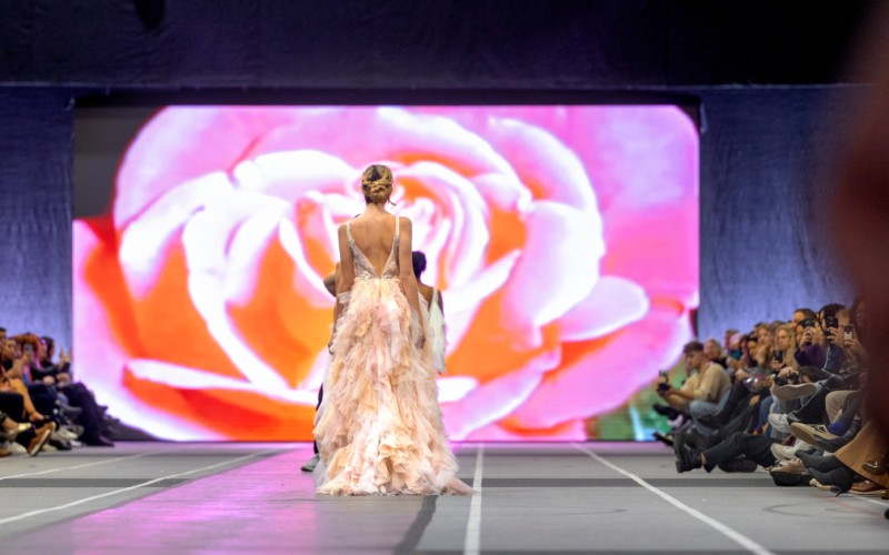 model in a pink gown walking towards a large screen with an open pink flower on it
