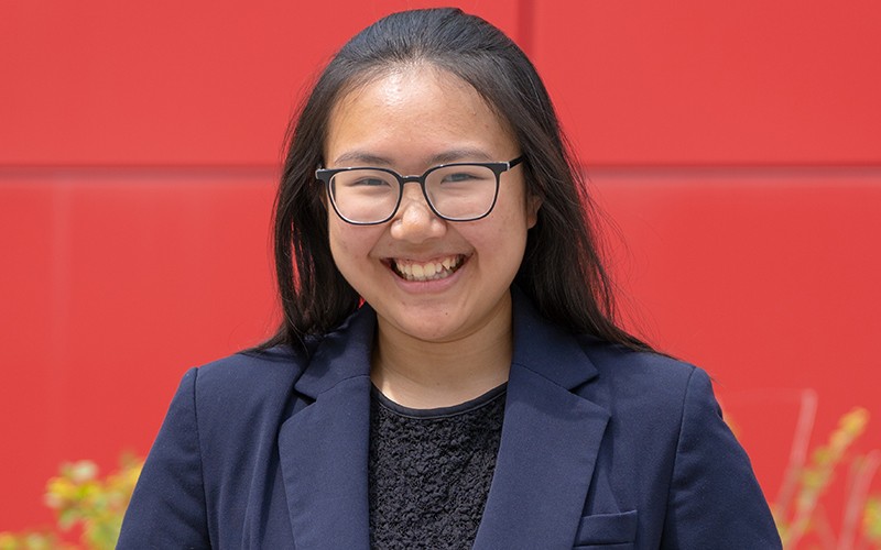 college senior wearing glasses and a blue blazer smiling with a red background