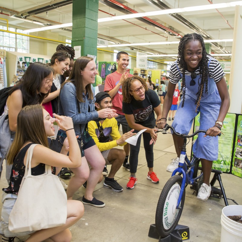 College student on a stationary bike powering a small appliance as fellow students look on and cheer